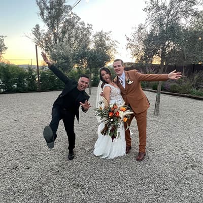 The DJ leaping beside a bride and groom on a gravel courtyard at golden hour